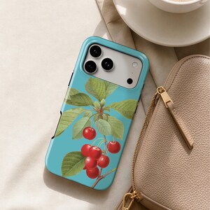 A teal-colored phone case with a cherry tree design, a white coffee cup, and a tan purse are arranged on a beige surface.