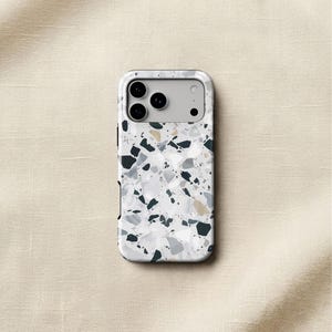 May include: A rectangular smartphone case featuring a white base with a multi-colored terrazzo pattern consisting of scattered black, dark gray, light gray, and tan stone-like fragments. The case has a cutout for a triple-lens camera system.