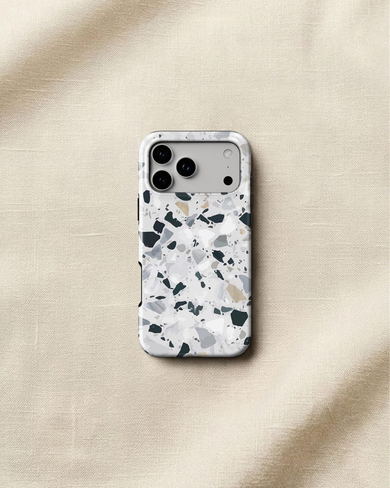 May include: A rectangular smartphone case featuring a white base with a multi-colored terrazzo pattern consisting of scattered black, dark gray, light gray, and tan stone-like fragments. The case has a cutout for a triple-lens camera system.