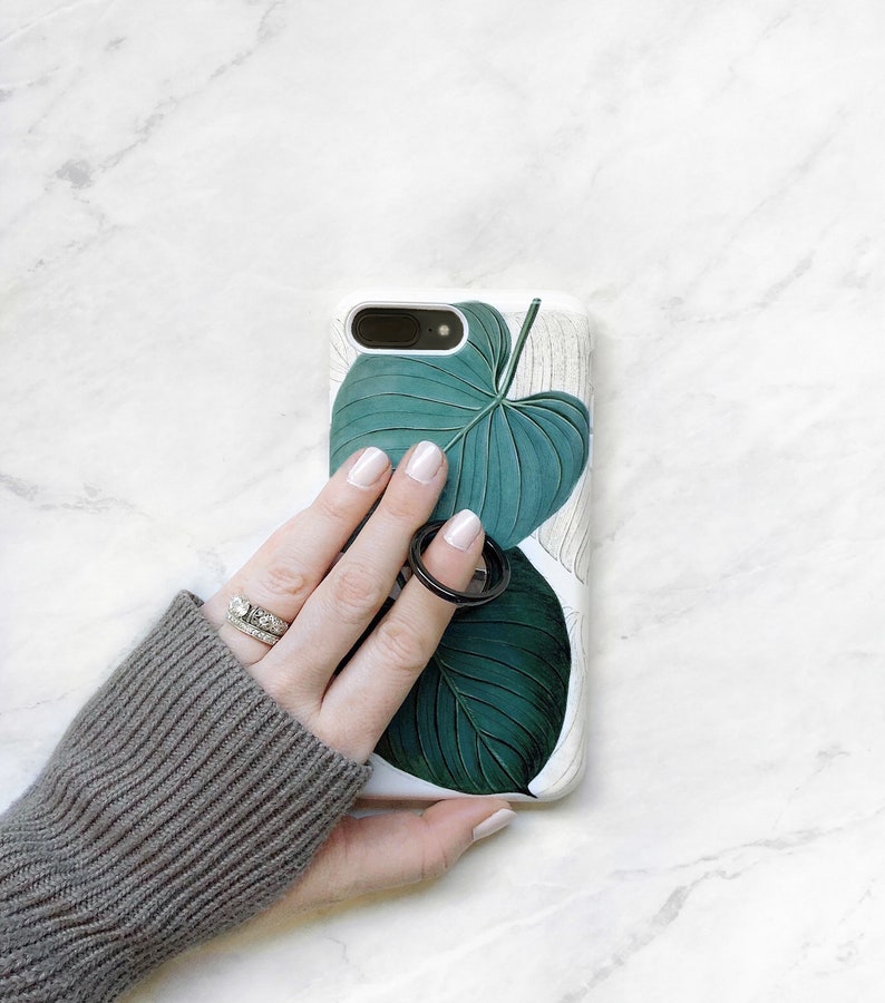 May include: A white phone case with a green tropical leaf design. A hand is holding the phone case and wearing two rings.