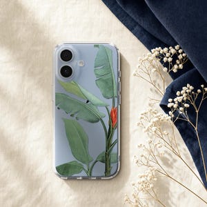 a clear phone case with a tropical leaf design, placed on a beige surface next to a blue fabric and some white flowers.