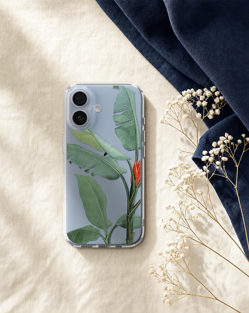 a clear phone case with a tropical leaf design, placed on a beige surface next to a blue fabric and some white flowers.