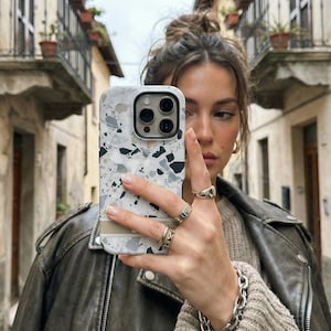 May include: A smartphone case featuring a white, gray, and black terrazzo pattern with a horizontal beige stripe across the lower section. The case has a cutout for a triple-lens camera system and is designed to fit a modern mobile device.
