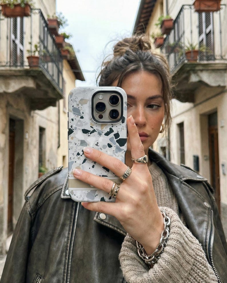 May include: A smartphone case featuring a white, gray, and black terrazzo pattern with a horizontal beige stripe across the lower section. The case has a cutout for a triple-lens camera system and is designed to fit a modern mobile device.
