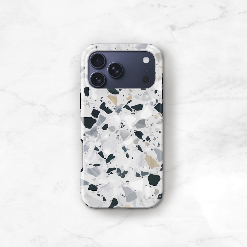 May include: A smartphone case featuring a light gray terrazzo pattern with scattered fragments in shades of white, charcoal black, and muted tan. The case includes a rectangular camera cutout at the top left, revealing three circular lenses.