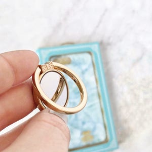 May include: A gold phone ring holder with a bow design. The ring is in a hand, and the background is a light blue and white surface.