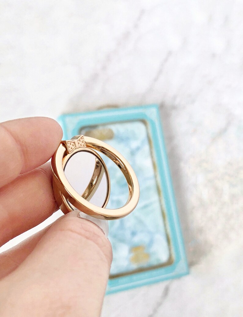 May include: A gold phone ring holder with a bow design. The ring is in a hand, and the background is a light blue and white surface.