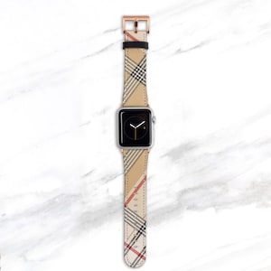May include: A silver Apple Watch with a beige, black, and red plaid band. The band has a rose gold buckle.