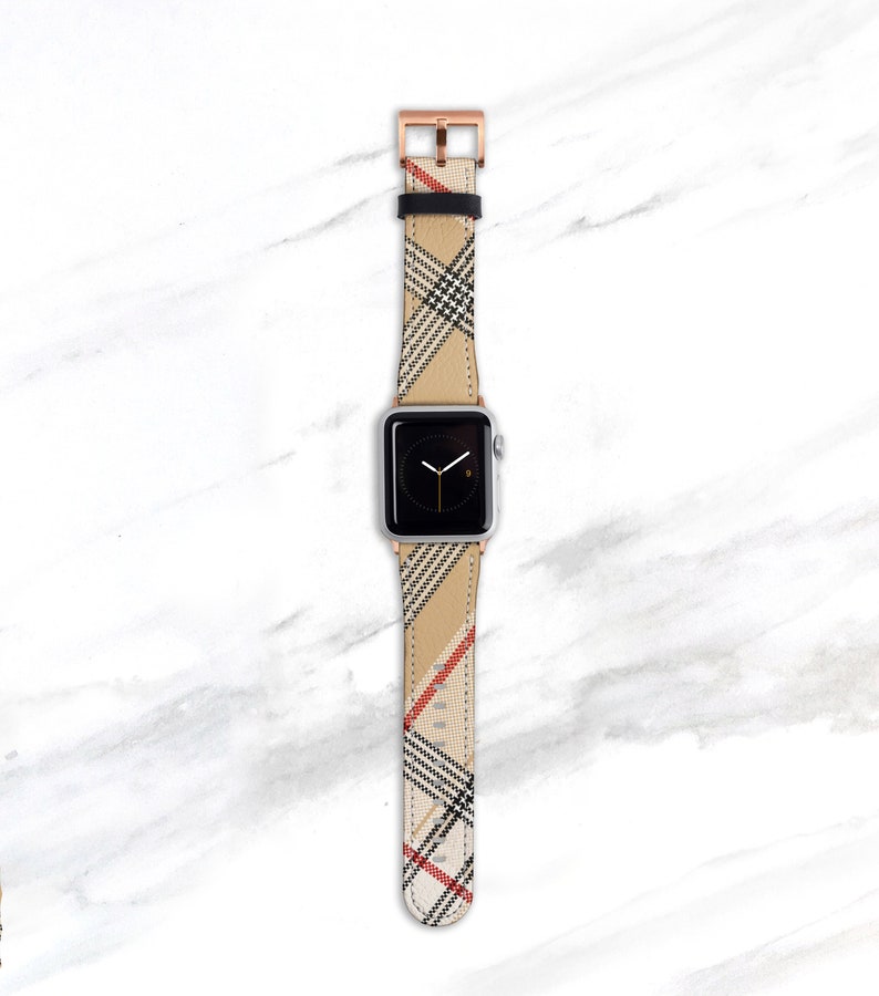 May include: A silver Apple Watch with a beige, black, and red plaid band. The band has a rose gold buckle.