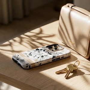 May include: A smartphone case featuring a white, gray, and black terrazzo pattern with a horizontal beige stripe across the bottom. The stripe contains the letters JMS in white. The case has a gold-toned camera cutout and black side buttons.