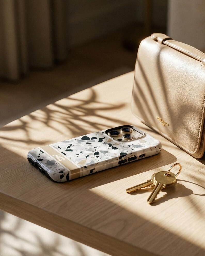 May include: A smartphone case featuring a white, gray, and black terrazzo pattern with a horizontal beige stripe across the bottom. The stripe contains the letters JMS in white. The case has a gold-toned camera cutout and black side buttons.