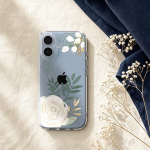 a clear phone case with a floral design, including a white rose and greenery, placed on a beige surface next to a blue fabric and some white flowers.