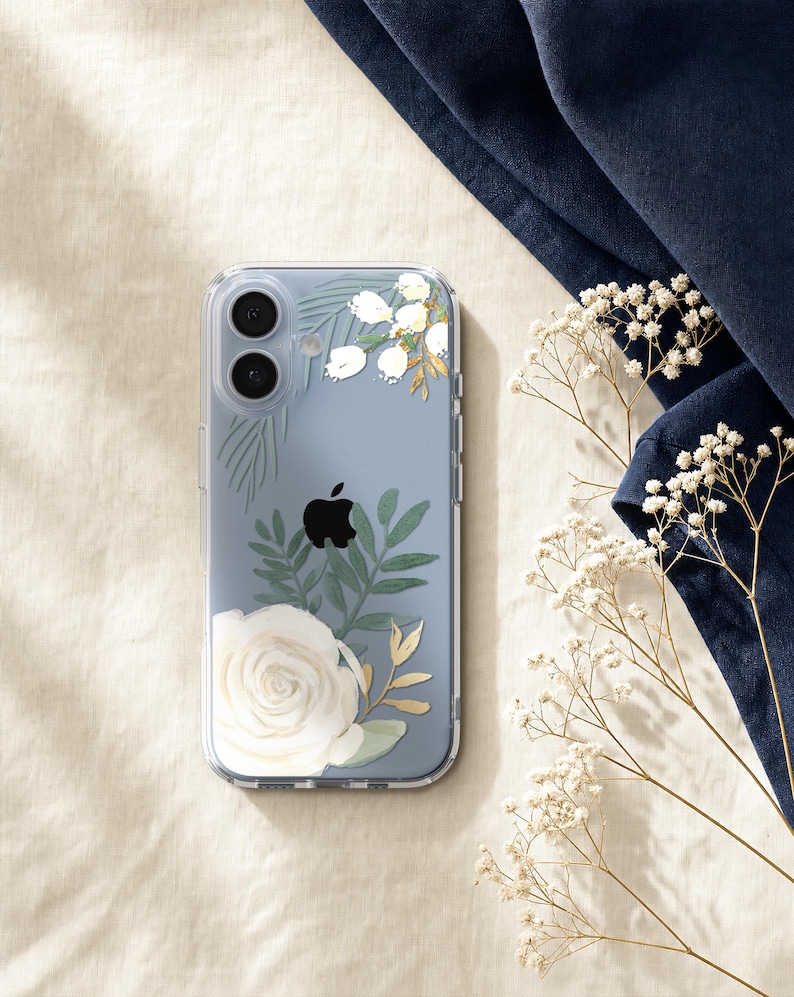 a clear phone case with a floral design, including a white rose and greenery, placed on a beige surface next to a blue fabric and some white flowers.