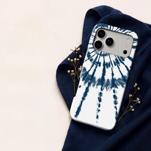 A white iPhone case with a tie-dye design is placed on a blue fabric, with small white flowers and a white cloth visible in the background.