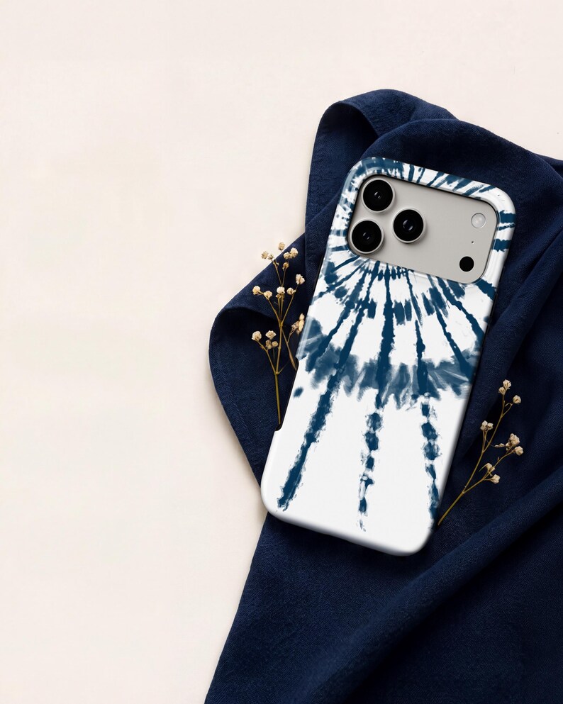 A white iPhone case with a tie-dye design is placed on a blue fabric, with small white flowers and a white cloth visible in the background.