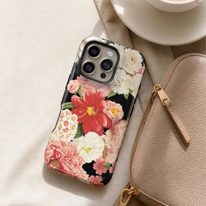 A floral phone case with a red and pink flower design rests on a white surface, accompanied by a cup of coffee and a beige purse.
