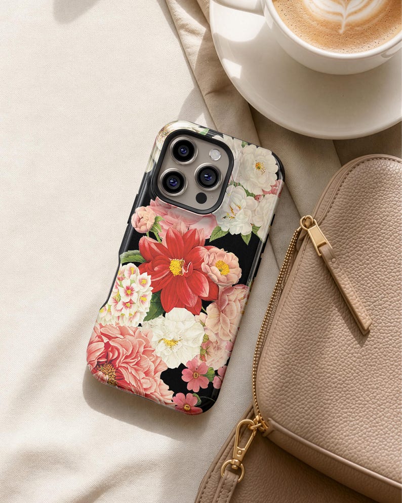 A floral phone case with a red and pink flower design rests on a white surface, accompanied by a cup of coffee and a beige purse.
