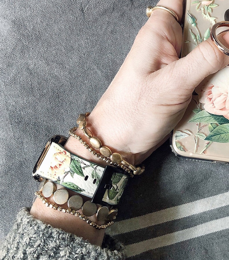 Op de afbeelding: Een wit en groen bloemenpatroon horlogebandje met een zwarte gesp. Het horlogebandje wordt gedragen op een pols met meerdere gouden kralen armbanden.