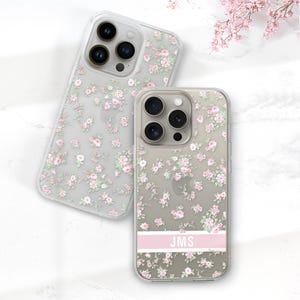 May include: Two clear phone cases with a floral pattern of small pink and white flowers. One case has a pink stripe with the initials "JMS". The cases are on a white surface with pink flowers in the background.