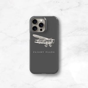 Peut inclure: Coque de t&eacute;l&eacute;phone gris fonc&eacute; avec une illustration blanche d'un avion vintage. Le texte "FLIGHT PLANS" est imprim&eacute; en dessous. Le bo&icirc;tier est dot&eacute; d'un protecteur d'objectif de cam&eacute;ra transparent.