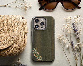 May include: A dark olive green smartphone case with a wood grain texture and a small floral illustration in the bottom left corner. The case features a cutout for a triple-lens camera system and is surrounded by a straw bag and sunglasses.