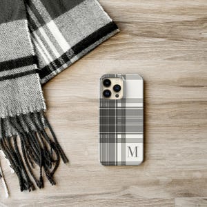 May include: A smartphone case featuring a black, white, and gray plaid pattern with a prominent letter M monogram in the lower right corner. The case has a cutout for a triple-lens camera system on the upper left side of the back panel.