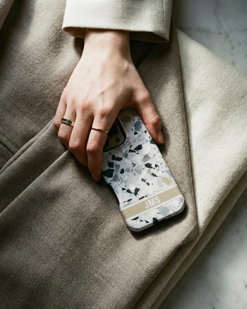 May include: A smartphone case featuring a white, gray, and black terrazzo pattern with a horizontal beige stripe across the lower section. The stripe contains the letters JMS printed in a clean, sans-serif font. The case has a smooth finish.