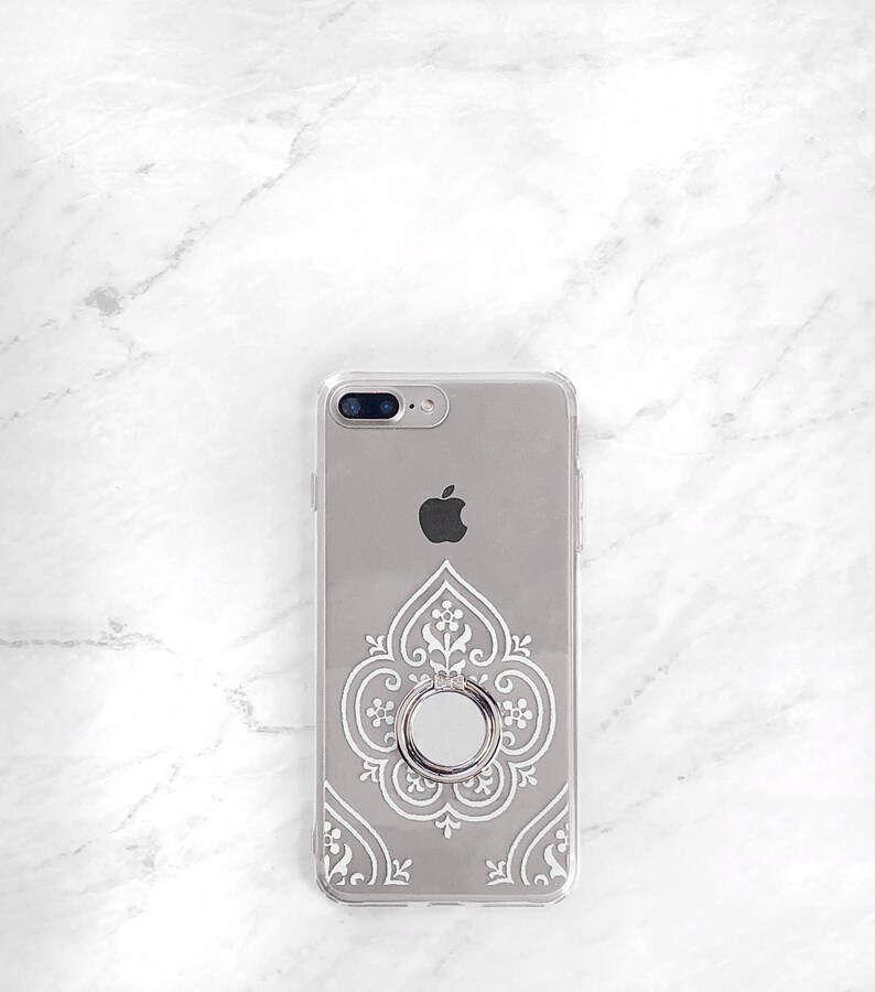 May include: A clear phone case with a white intricate design and a silver ring holder. The case is designed for an iPhone.