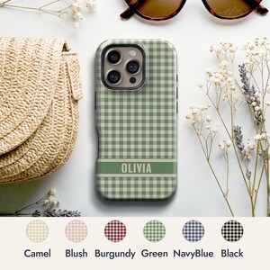 May include: A sage green and white gingham patterned phone case featuring a horizontal band with the name OLIVIA printed in white. The case has a camera cutout and is displayed alongside a straw bag, sunglasses, and dried flowers.