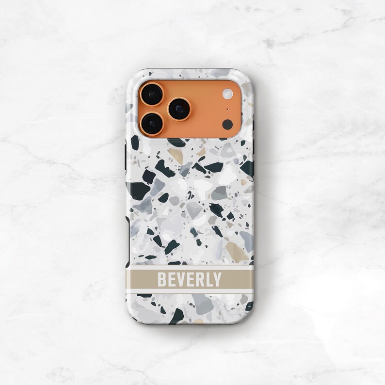 May include: A rectangular phone case featuring a white, gray, and black terrazzo pattern. The upper section has an orange camera cutout area. A horizontal beige band across the lower portion displays the name BEVERLY in white capital letters.