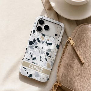 May include: A smartphone case featuring a white, gray, and black terrazzo pattern with a horizontal beige band across the lower section. The name PARKER is printed in white capital letters on the beige band. The case has a cutout for a triple camera.