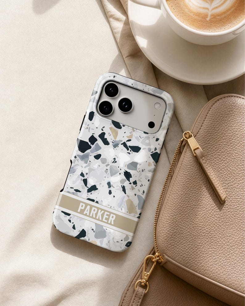 May include: A smartphone case featuring a white, gray, and black terrazzo pattern with a horizontal beige band across the lower section. The name PARKER is printed in white capital letters on the beige band. The case has a cutout for a triple camera.