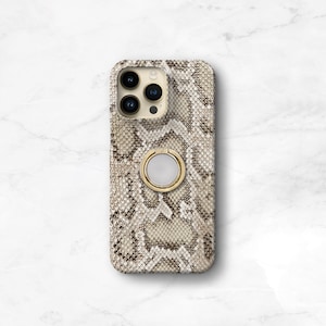 May include: A phone case with a snakeskin pattern in shades of brown and beige. The case has a gold-colored ring holder on the back. The phone case has a camera cutout with three lenses.
