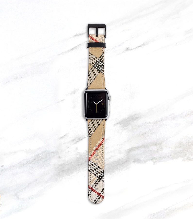 May include: A silver Apple Watch with a black and beige plaid patterned band. The band has red accents.