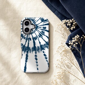 a white phone case with a tie-dye pattern, placed on a beige surface next to some white flowers and a blue fabric.