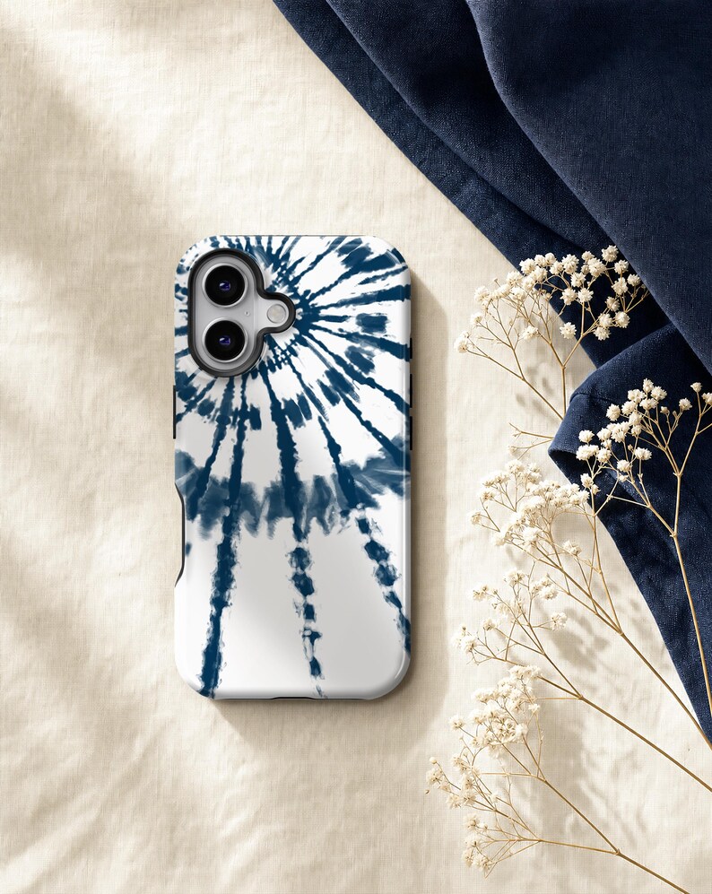a white phone case with a tie-dye pattern, placed on a beige surface next to some white flowers and a blue fabric.