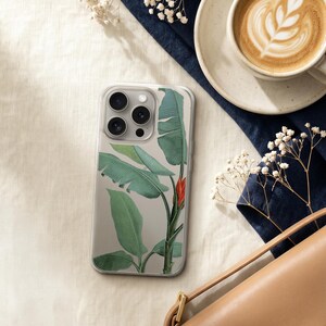 a smartphone with a plant-themed case, a cup of coffee, a purse, and some white flowers on a white surface.