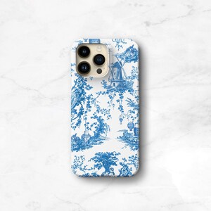 May include: A white smartphone case featuring a blue toile de jouy pattern. The design displays intricate illustrations of a windmill, trees, foliage, and a hot air balloon, creating a detailed landscape scene across the entire surface.