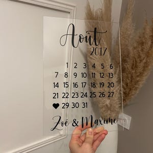 May include: A clear acrylic calendar with the month of August 2017 printed on it. The calendar has a black heart marking the 29th and the text "Zoé & Maxime" at the bottom.