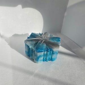 May include: A decorative, square resin box with a lid. The box is a gradient of blue, white, and silver, with a glittery finish. The box has a textured, geometric design and is sitting on a white surface.