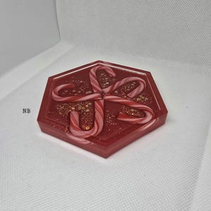 Könnte beinhalten: Ein roter sechseckiger Untersetzer mit einem Zuckerstangen-Design. Der Untersetzer ist mit rotem Harz und goldenem Glitzer gefüllt. Vier Zuckerstangen sind in der Mitte angeordnet und schaffen ein festliches Urlaubs-Thema.