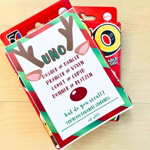 May include: A festive UNO card game box with a custom reindeer-themed design. The card features reindeer antlers, ears, and the names of Santa's reindeer. Text on the card reads "but do you recall? You're my favourite students of all!"