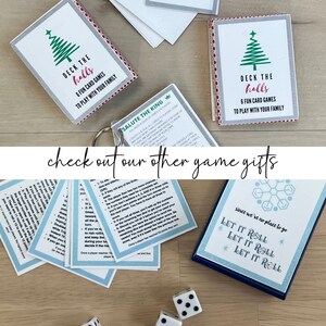May include: Various card game sets with festive designs are arranged on a wooden surface. One set features "Deck the Halls" and a Christmas tree graphic. Another set has the text "Let it Roll". Dice are scattered nearby. The phrase "check out our other game gifts" is also visible.