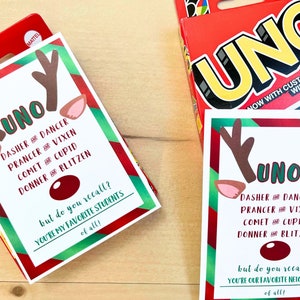 May include: Two UNO card game boxes and cards with a reindeer theme. The cards feature reindeer antlers, ears, and a red nose, with text that reads "UNO" and the names of Santa's reindeer. The box is red with the word "UNO" in large yellow letters.