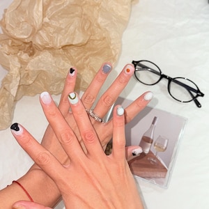 Puede incluir: Primer plano de manos con uñas pintadas. Las uñas están pintadas con diferentes colores y diseños, incluyendo blanco, gris, negro y azul claro con un acento dorado.