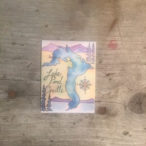 May include: A watercolor illustration of Lake Pend Oreille, a lake in Idaho, with a compass rose and the words "Lake Pend Oreille" written in cursive.