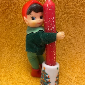 May include: A vintage Christmas elf figurine holding a red candle with glitter. The elf is dressed in a red hat, green outfit, and red boots. The candle is in a white ceramic holder decorated with Christmas trees and candles.