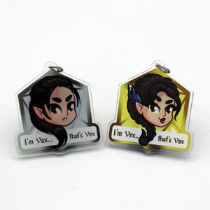 May include: Two acrylic keychains featuring cartoon illustrations of a female elf with dark hair. One keychain is silver with the text "I'm Vax... that's Vex". The other keychain is gold with the text "I'm Vex... that's Vax".