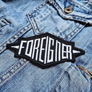 Op de afbeelding: Zwart-witte geborduurde patch met het woord "FOREIGNER" in wit op een zwarte achtergrond. De patch heeft de vorm van een ruit met puntige randen.