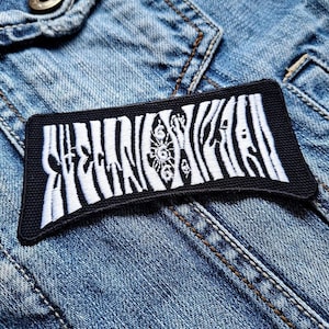 Pode incluir: Um patch retangular bordado em preto e branco com a palavra "Zebra" em uma fonte estilizada. O patch apresenta um padrão de listras de zebra e é colocado em uma jaqueta jeans.